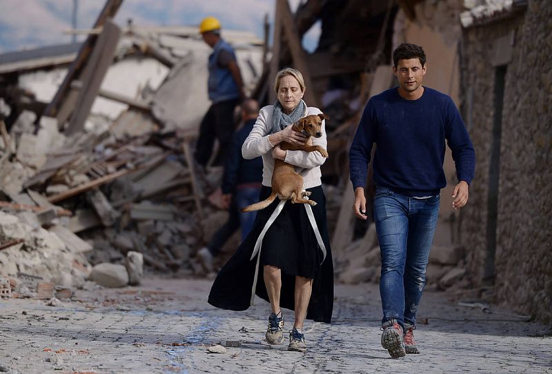 Una mujer sostiene un perro en sus brazos mientras camina junto a un hombre entre los edificios derruidos en Amatrice