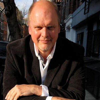 Retrato de Michael Daugherty, compositor, con americana oscura y camisa blanca, en una calle soleada con coches y casas al fondo.