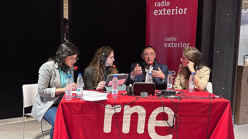 Cuatro personas (dos hombres, dos mujeres) en una mesa de radio durante una emisión.  Micrófonos, papeles y un mantel rojo con el logo de RNE son visibles.  Conversación activa.