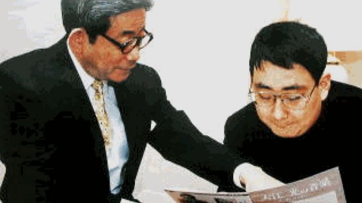 Padre e hijo sentados, el mayor señala una publicación titulada \"ASC News Post\". Ambos llevan gafas; uno viste traje oscuro y el otro camisa oscura.