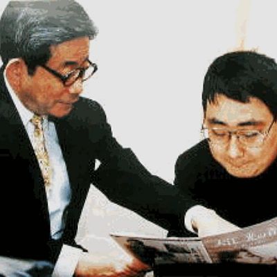 Padre e hijo sentados, el mayor señala una publicación titulada \"ASC News Post\". Ambos llevan gafas; uno viste traje oscuro y el otro camisa oscura.
