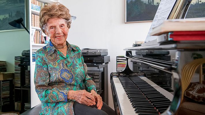 Mujer mayor sonriente junto a un piano con partituras; al fondo, libros y equipo de música. Ambiente cálido y acogedor.