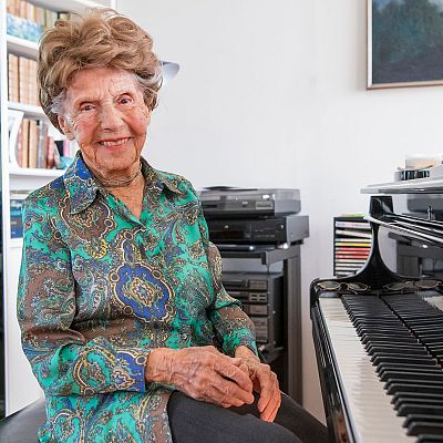 Mujer mayor sonriente junto a un piano con partituras; al fondo, libros y equipo de música. Ambiente cálido y acogedor.