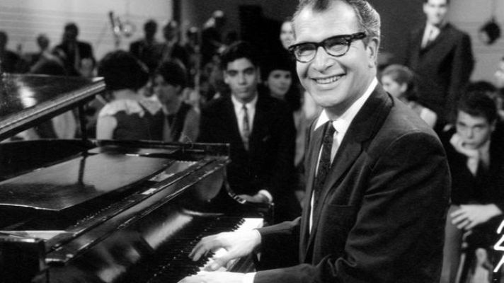 Dave Brubeck, con traje y gafas, sonríe mientras toca un piano de cola ante un público. Foto en blanco y negro.