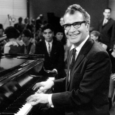 Dave Brubeck, con traje y gafas, sonríe mientras toca un piano de cola ante un público. Foto en blanco y negro.
