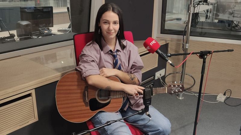 Joven con guitarra acústica en estudio de radio, micrófono con logo 'rne me', camisa malva y corbata a rayas.  Expresión relajada.