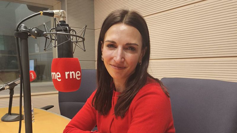 Mujer con jersey rojo en estudio de radio RNE, frente a micrófono con funda roja y logo blanco. Expresión serena. Posible grabación de programa.