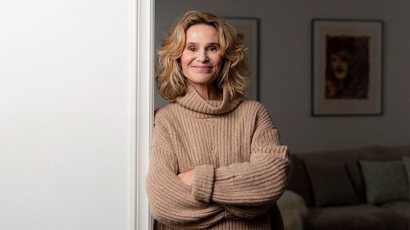 Mujer rubia de mediana edad, sonriente, apoyada en puerta blanca, jersey de punto grueso, sala de estar con sofá y cuadros.