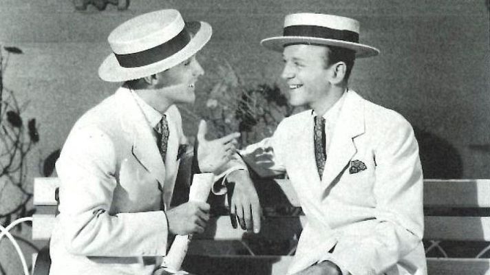Gene Kelly y Fred Astaire, en trajes blancos y sombreros de paja, conversan animadamente sentados.  Imagen en blanco y negro con fondo de estudio.