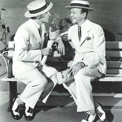 Gene Kelly y Fred Astaire, en trajes blancos y sombreros de paja, conversan animadamente sentados.  Imagen en blanco y negro con fondo de estudio.