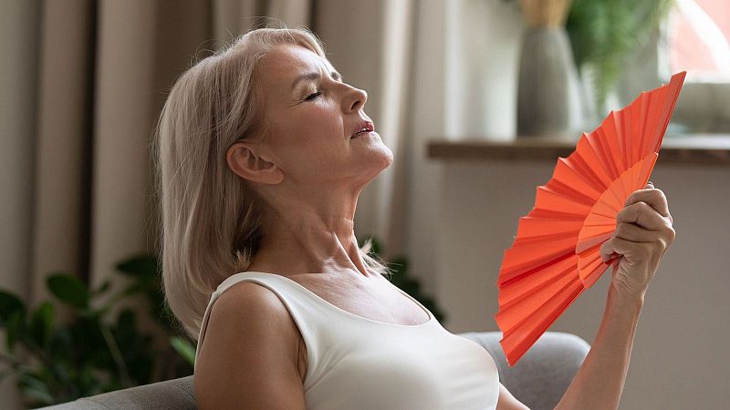 Mujer mayor abanicándose con un abanico naranja sentada en un sofá.  Expresión de calor. Fondo con planta y cortina.