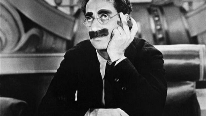Groucho Marx, en blanco y negro, sentado con gafas redondas y bigote, muestra una expresión pensativa en un ambiente clásico.