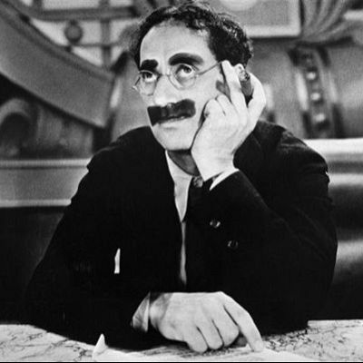 Groucho Marx, en blanco y negro, sentado con gafas redondas y bigote, muestra una expresión pensativa en un ambiente clásico.