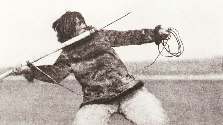 Nanook, con ropa de piel, lanza un arpón de madera con una cuerda en la mano.  Imagen en blanco y negro que evoca la vida inuit.