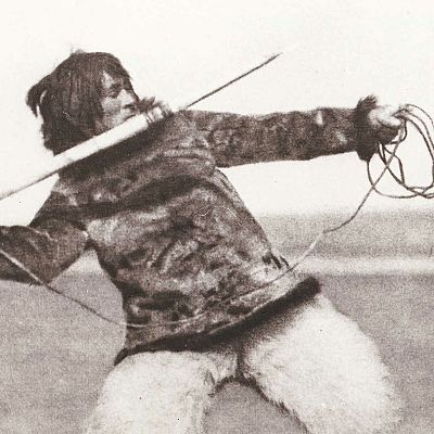 Nanook, con ropa de piel, lanza un arpón de madera con una cuerda en la mano.  Imagen en blanco y negro que evoca la vida inuit.