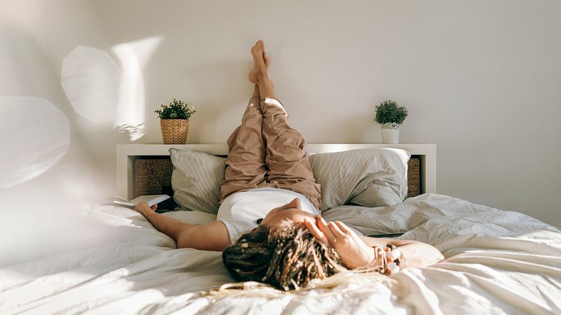 Mujer descansando en cama, con móvil en mano y pelo en trenzas. Lleva camiseta blanca y pantalones beige. Ambiente tranquilo y luminoso.