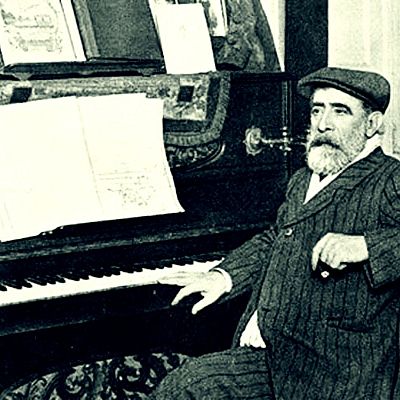 Tomás Bretón, compositor español, sentado al piano con partituras.  Viste traje a rayas y gorra; la imagen es en blanco y negro.