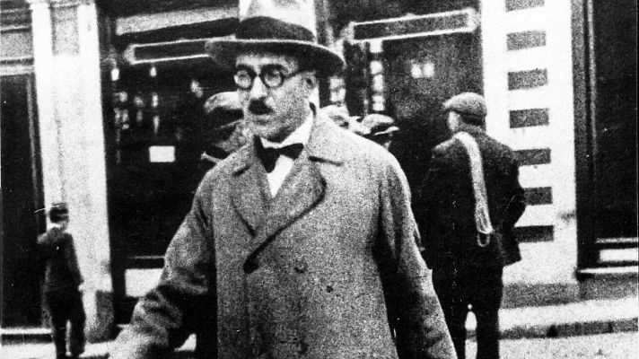 Fotografía antigua en blanco y negro de Fernando Pessoa en la calle, con sombrero, gafas y abrigo; fondo con edificios y personas borrosas.
