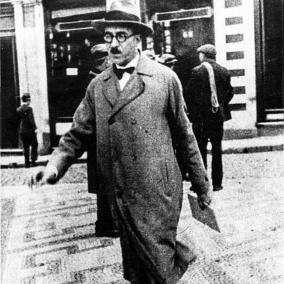 Fotografía antigua en blanco y negro de Fernando Pessoa en la calle, con sombrero, gafas y abrigo; fondo con edificios y personas borrosas.