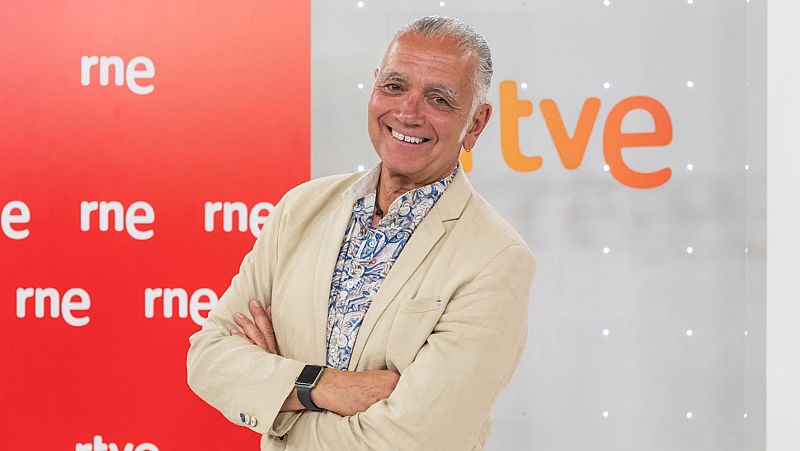 JR Lucas posa sonriente con americana beige y camisa estampada, ante un fondo con los logotipos de RNE y TVE.