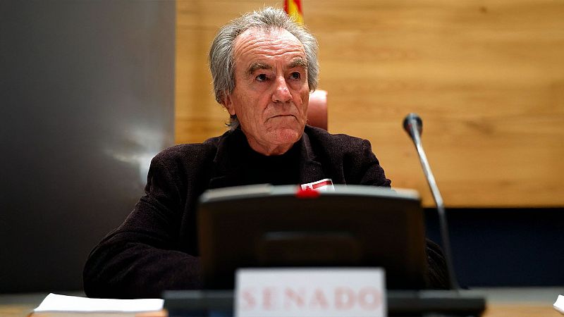 Javier Pérez Royo, con chaqueta oscura y camisa negra, se encuentra sentado frente a un micrófono en el Senado, con un letrero que lo identifica. El fondo muestra una bandera española.
