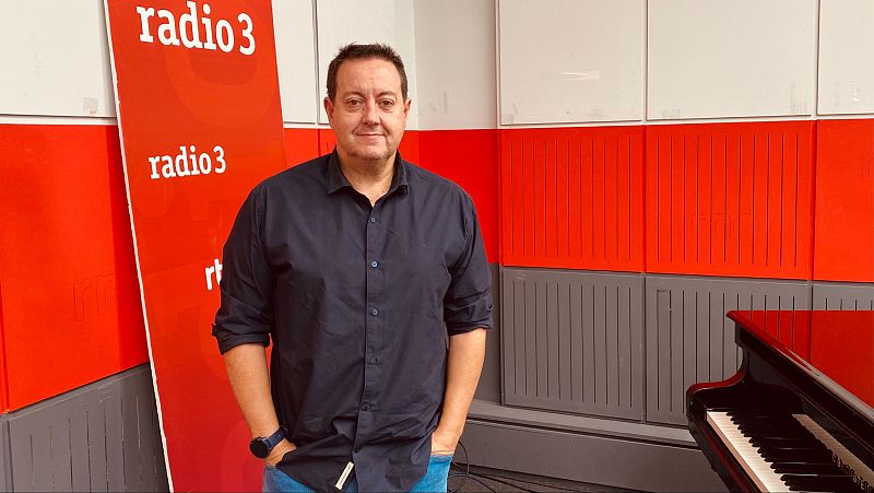Antoni Daimiel, con camisa azul oscura, posa en un estudio frente a un cartel y un piano de cola. El fondo presenta una pared dividida en secciones de colores.