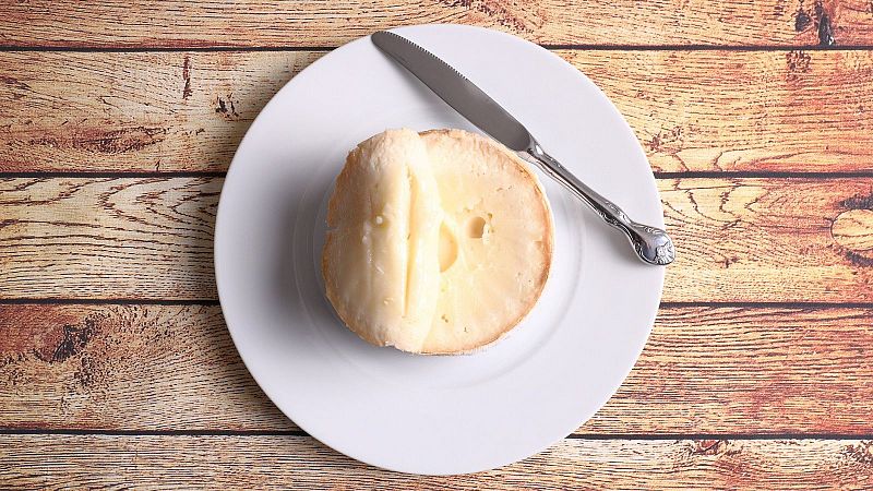 Una Torta del Casar, con un corte que revela su interior cremoso, se presenta en un plato blanco junto a un cuchillo decorado, sobre una mesa de madera.