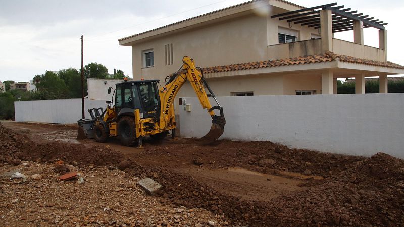 Una excavadora amarilla de la marca New Holland opera en un terreno, con una casa de dos pisos color beige al fondo, que tiene un balcón con estructura de madera y una valla blanca.