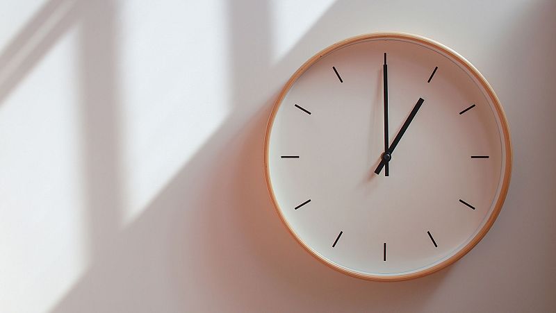 Un reloj de pared con marco de madera clara marca la una en punto, con manecillas negras sobre una esfera blanca. El fondo es una pared blanca con sombras.