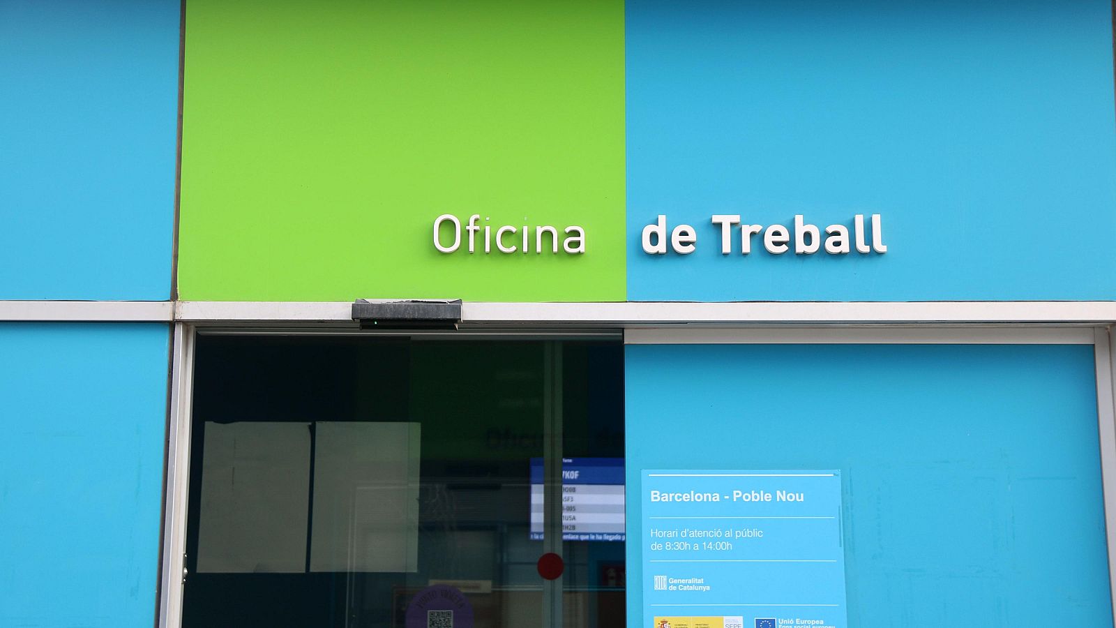 En la fachada de la oficina, se observa un cartel informativo con el horario de atención al público y la ubicación en Barcelona, junto a los logotipos de la Generalitat de Catalunya y la Unión Europea.