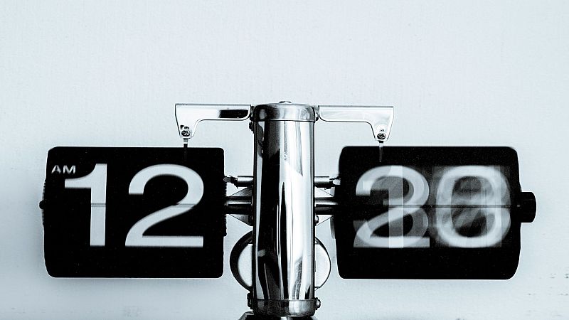 Un reloj de solapa plateado de estilo retro muestra la hora en dos paneles negros: "12" y "20", indicando las 12:20 AM. El panel izquierdo indica "AM".