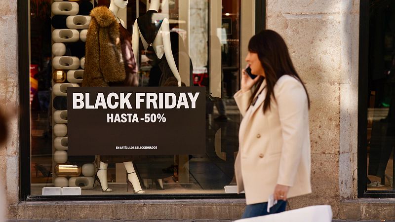 Un escaparate de una tienda de ropa anuncia el Black Friday con un cartel que promociona descuentos. Se exhiben prendas en maniquíes, mientras una mujer camina frente al escaparate hablando por teléfono y con una bolsa de compra.