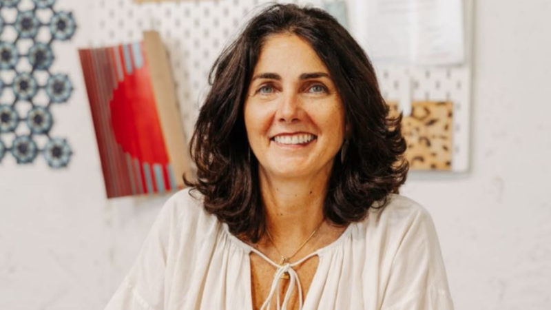 Una mujer con cabello castaño oscuro y ojos azules sonríe, vestida con una blusa blanca. El fondo muestra elementos decorativos en la pared, incluyendo un mosaico y una lámina con diseños.