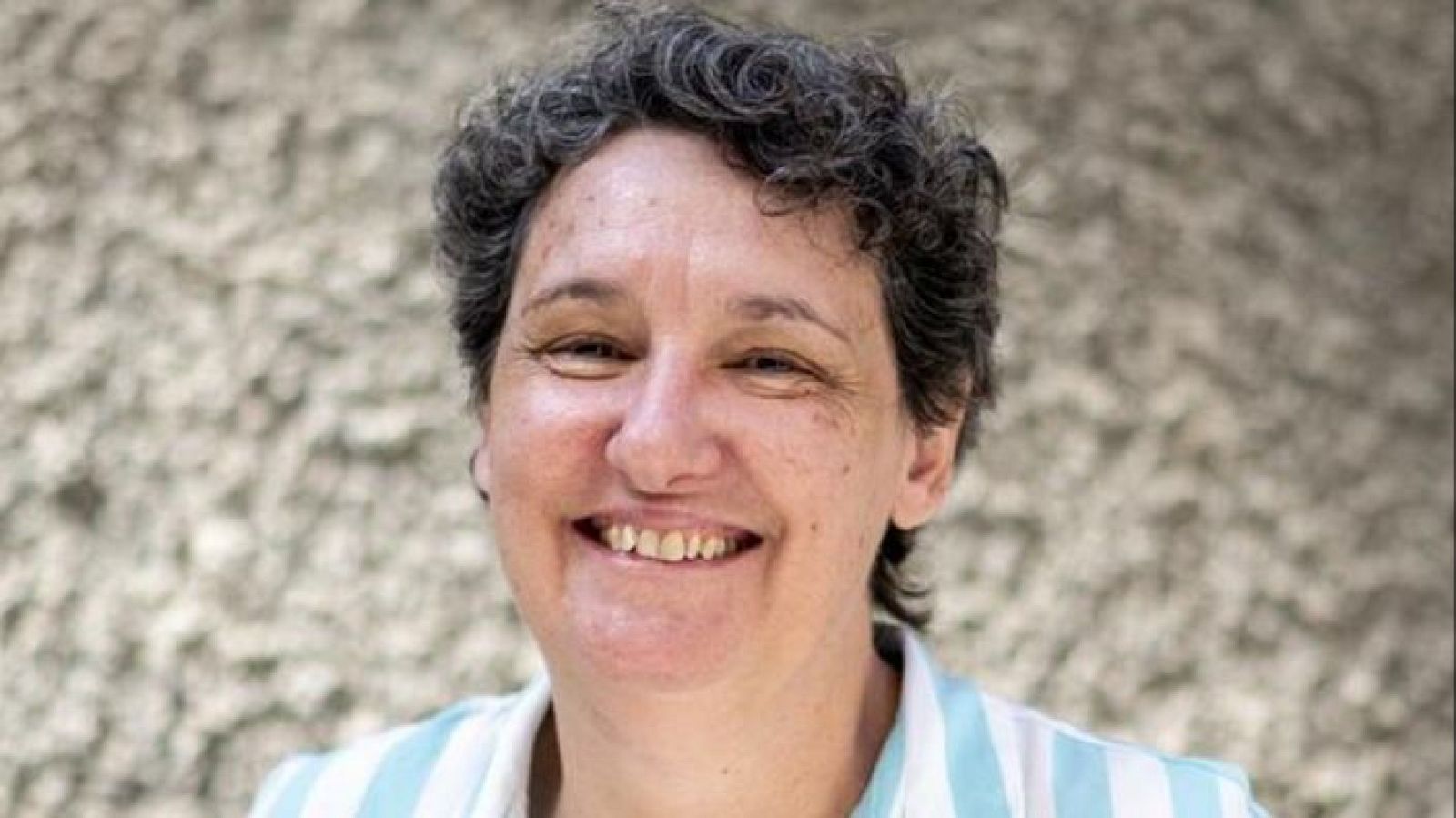 Retrato de Beatriz Gimeno, sonriente, con pelo corto y rizado gris oscuro, vestida con camisa de rayas azules y blancas. Fondo desenfocado.