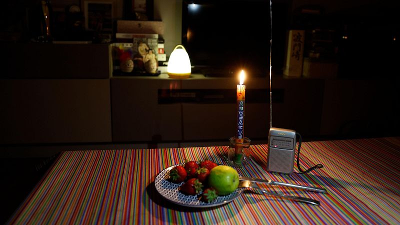 Escena a la luz de vela con frutas, cubiertos y radio portátil, en un interior con TV apagada y lámpara tenue, evocando un apagón.