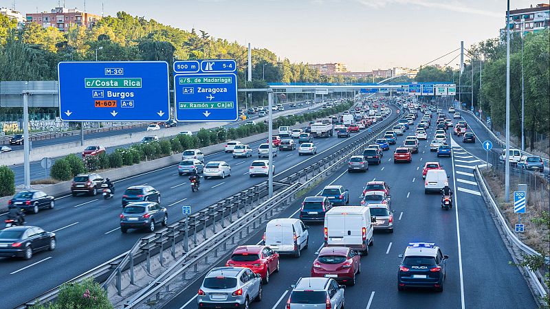 Vista aérea de autopista congestionada en Madrid al atardecer, con tráfico denso, coches, furgonetas, motos y un coche de policía con luces de emergencia. Entorno urbano con vegetación y edificios.