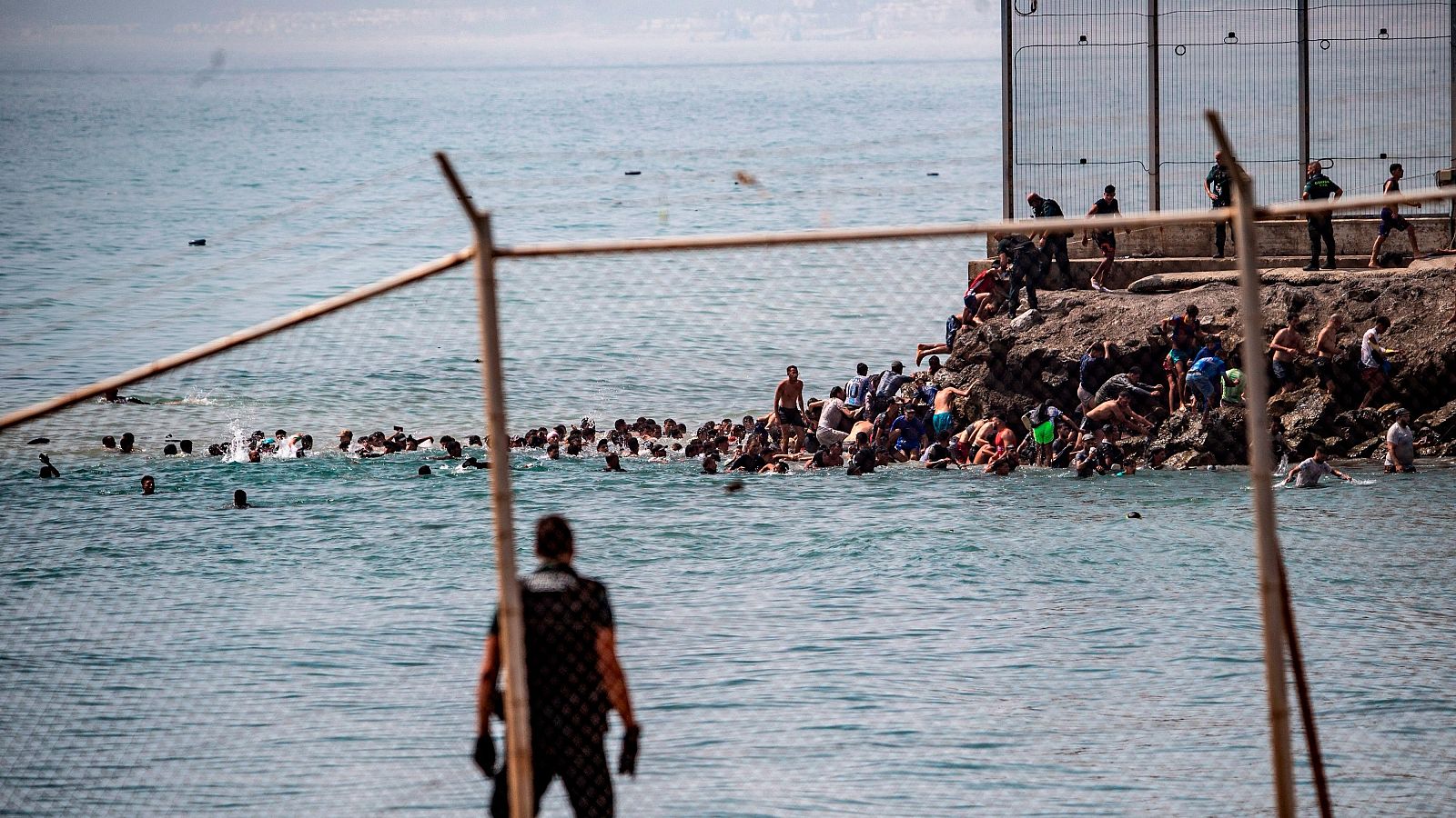 Multitud cruza frontera marítima en Ceuta, España. Personas nadan y trepan rocas hacia una valla, mientras agentes observan.