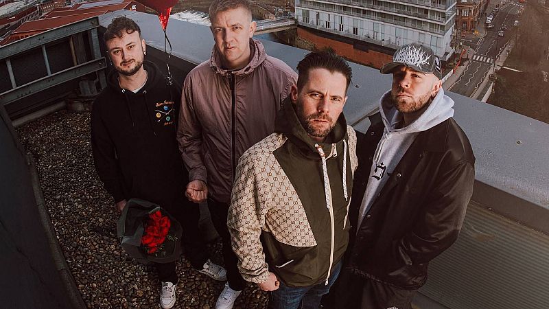 Cuatro hombres de la banda The Reytons posan en una azotea urbana con un paisaje de edificios al fondo. Destacan sus atuendos variados y accesorios como un ramo de rosas y un globo en forma de corazón.