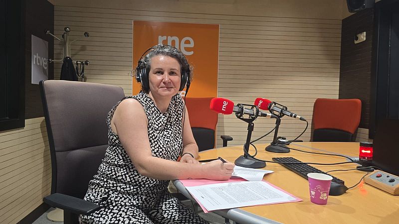 Lidia de la Iglesia, presentadora de RNE, en un estudio de radio con micrófonos, teclado y papeles. Viste blusa de puntos y auriculares.
