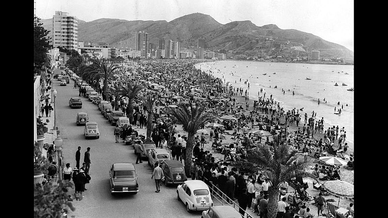 Escena de playa y paseo marítimo en España, años 60/70. Coches clásicos, miles de bañistas y sombrillas cubren la arena y el mar. Edificios y montañas al fondo.
