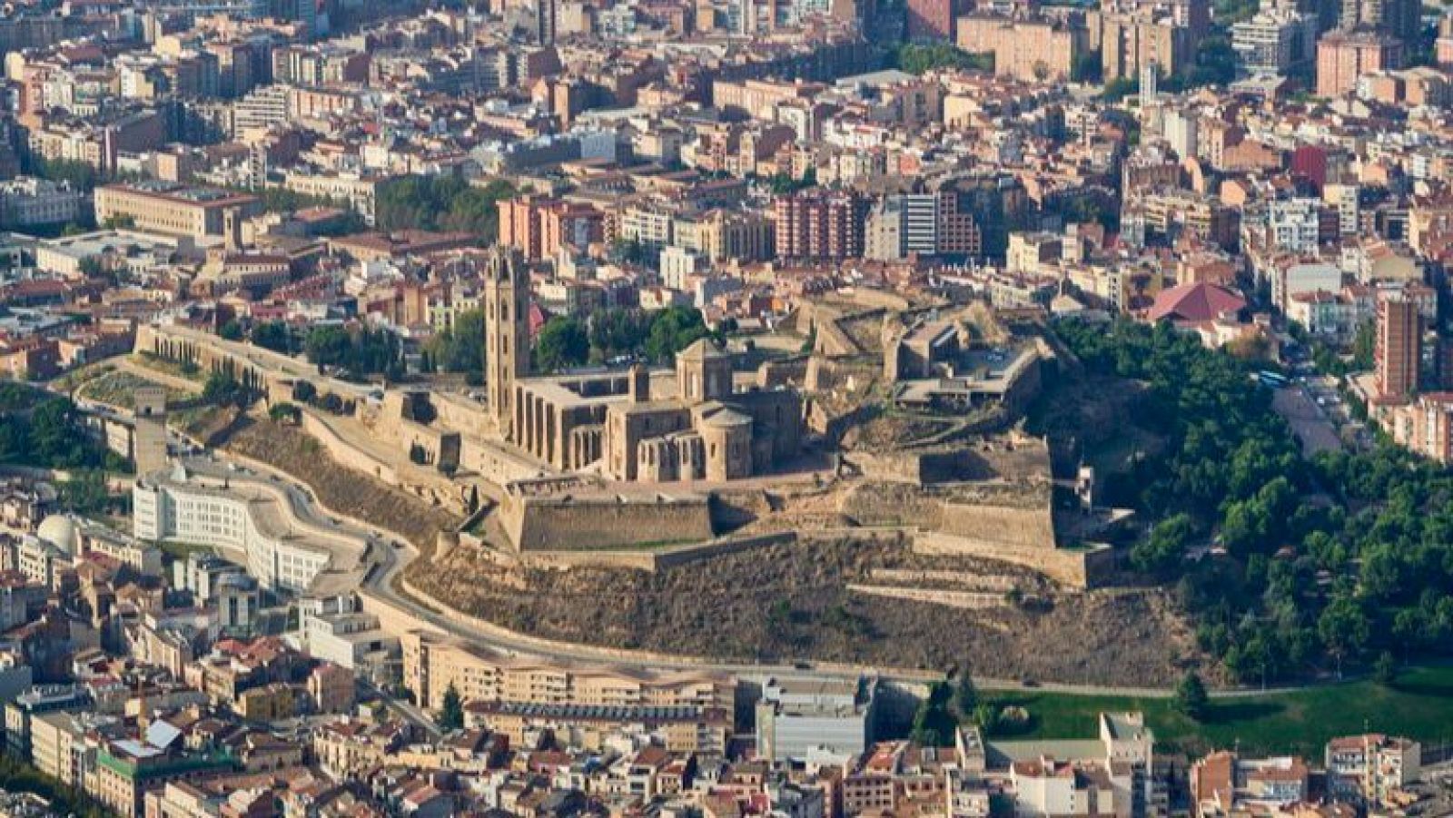 Vista aérea de Lleida, Cataluña, España, con la Seu Vella como punto focal. La histórica catedral y sus murallas dominan la colina, rodeadas por una extensa ciudad y áreas verdes.