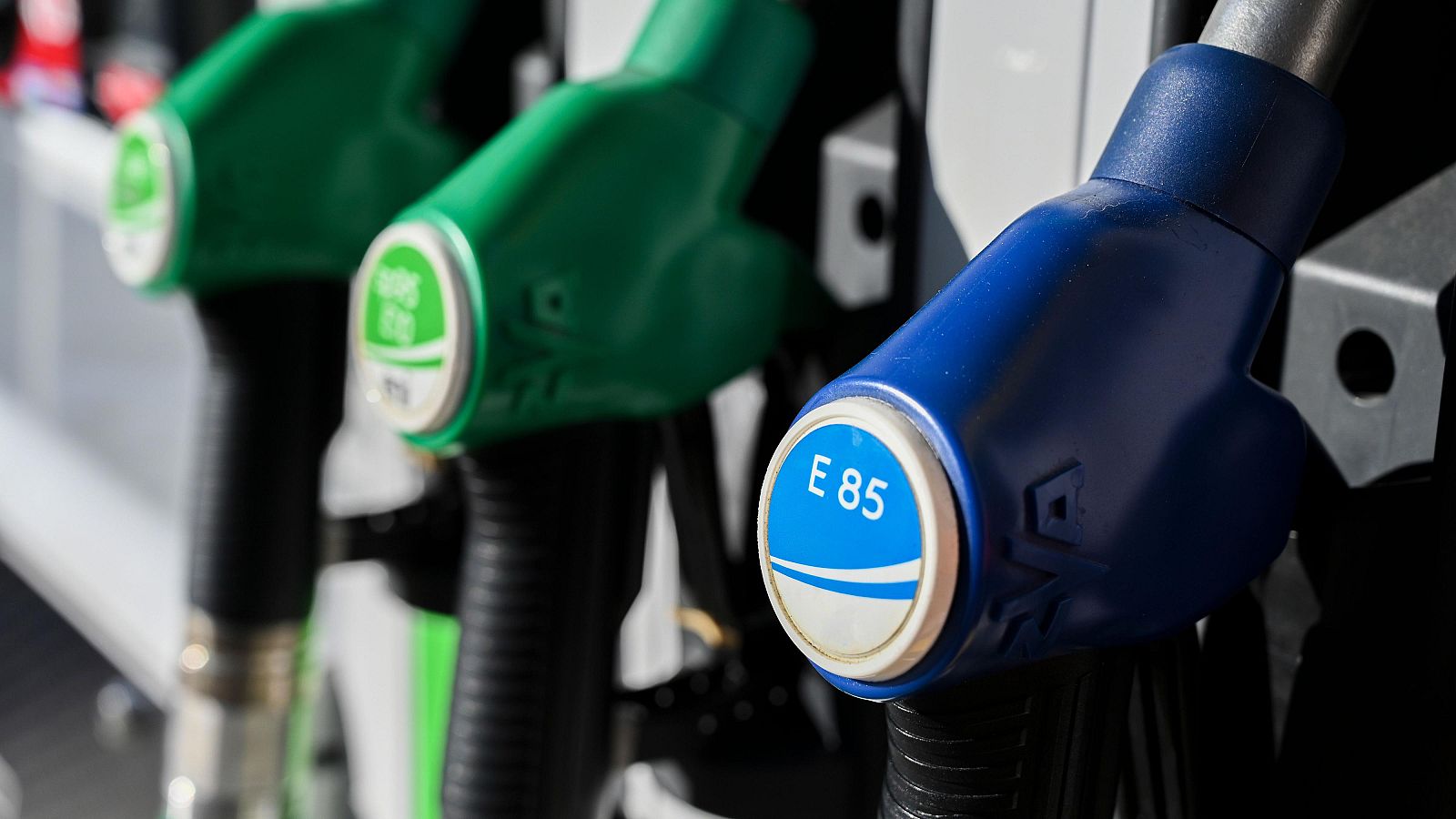Primer plano de tres boquillas de surtidor: una azul "E 85" y dos verdes "E10 95", dispuestas en diagonal. Destacan sus etiquetas y empuñaduras.