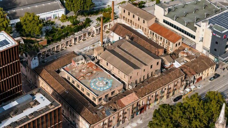 Vista aèria de La Escocesa, antiga fàbrica reconvertida en centre artístic a Barcelona, amb xemeneies de maó i un mosaic circular a un teulat.