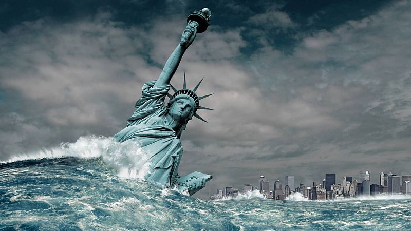 Estatua de la Libertad parcialmente sumergida en aguas turbulentas, con una ciudad al fondo bajo un cielo sombrío. La antorcha se mantiene encendida.