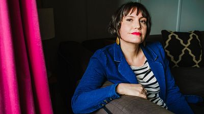 Retrato de Laia Jufresa, escritora mexicana, con chaqueta azul, camiseta de rayas y pendientes dorados, frente a una cortina fucsia y un sofá.