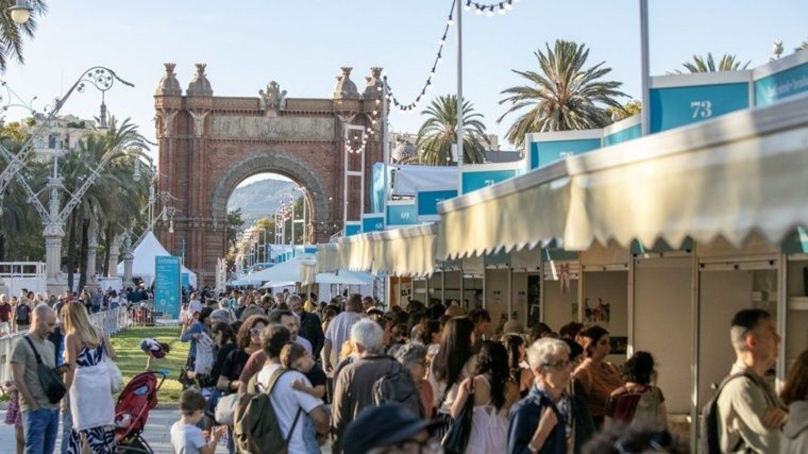 Vista panorámica de la Setmana del Llibre Català en el Passeig de Lluís Companys, Barcelona, con el Arc de Triomf al fondo. Multitud pasea entre puestos de exposición con toldos crema y azul.