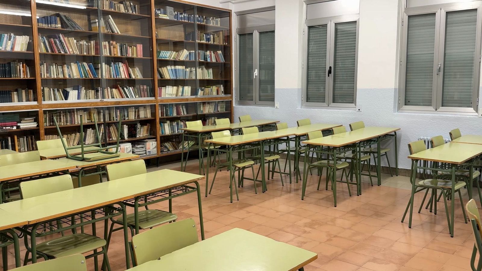 Aula vacía con estanterías de libros, mesas y sillas. Una silla volcada sobre una mesa. Paredes bicolor, ventanas con persianas bajadas y suelo de baldosas.