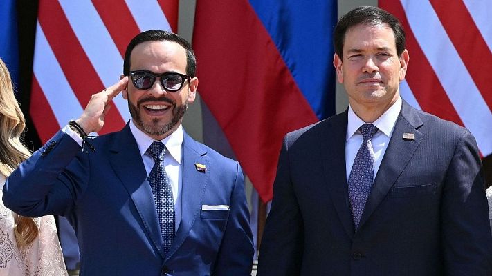 Juan Guaidó saluda militarmente con una sonrisa, vistiendo traje azul y pin de Venezuela. A su lado, Marco Rubio con traje oscuro y pin de EE.UU. Banderas de ambos países al fondo.