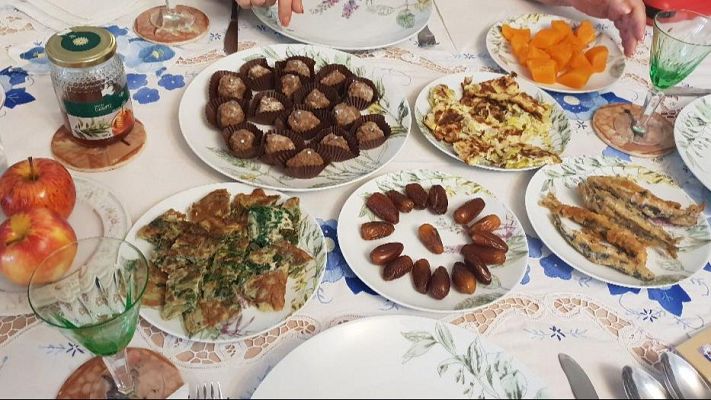 Mesa festiva con mantel floral azul y blanco, exhibiendo platos de calabaza, pescados fritos, dátiles, tortilla verde, dulces y miel, junto a copas verdes.