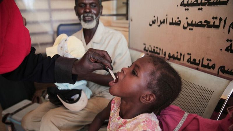 Niña recibe sustancia oral en centro de salud sudanés. Texto en árabe: 'Departamento de Inmunización contra enfermedades', 'Seguimiento del Crecimiento y la Nutrición'.